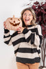 A woman wearing a black and cream striped knit sweater holds a carved jack-o'-lantern pumpkin, smiling and looking upward. She is standing indoors against a white wall with a decorative plant in the background.