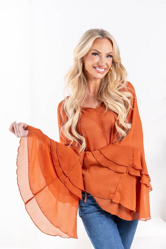 A woman wearing a rust orange long-sleeve blouse with a V-neck and tiered bell sleeve flared cuffs.