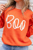 A close-up of a woman wearing a bright orange crewneck sweater with the word 'Boo' written across the front in large white cursive letters, paired with blue jeans.
