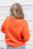 A woman from behind wearing a bright orange crewneck sweater paired with blue jeans.