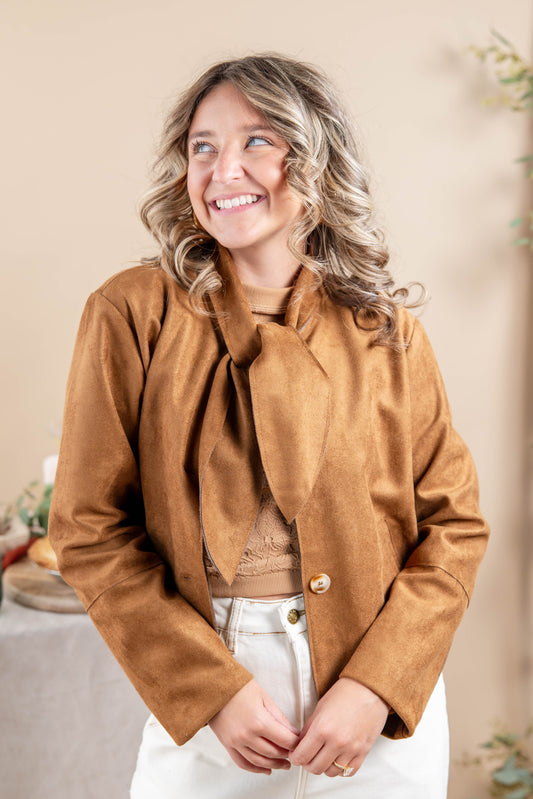 A woman wearing a cropped tan faux-suede jacket with a large bow tie neckline, styled over a beige top and white pants, posing against a warm beige background.
