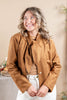 A woman wearing a cropped tan faux-suede jacket with a large bow tie neckline, styled over a beige top and white pants, posing against a warm beige background.