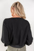 A woman viewed from behind wearing a black long-sleeve top with a wide boat neckline and gathered cuffs, paired with olive green pants, against a plain white studio background.