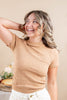 A woman wearing a beige textured short-sleeve mock neck top and white pants, smiling and looking upward in a warm indoor setting.