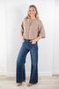 A woman modeling a beige short-sleeve button-front knit top and dark blue wide-leg jeans, standing in a bright indoor room with a white wall and light wood floor.