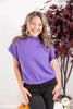 A smiling woman wearing a purple short-sleeve mock neck top and black pants, standing in front of a white wall with a decorative plant and pumpkins in the background.