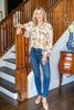 A woman stands in a home interior by a wooden staircase, wearing a long-sleeve button-up blouse with a beige, green, and orange abstract print, blue jeans, and beige pointed flats.