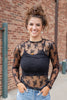 A woman wearing a black sheer lace long-sleeve top over a black ribbed camisole, paired with blue jeans and halloween themed jewelry.