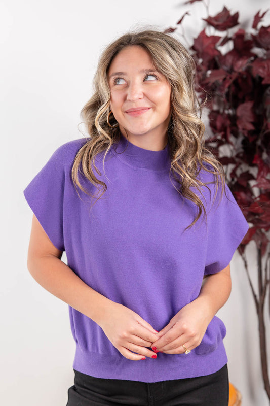 A woman with wavy light brown hair wearing a purple short-sleeve mock neck sweater, standing against a white wall with a decorative plant in the background.