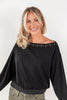 A smiling blonde woman wearing a black off-the-shoulder long-sleeve top with silver eyelet embellishments along the neckline, paired with olive green pants, against a plain white studio background.