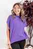 A woman wearing a purple cap dolman sleeve mock neck knit top and black pants, smiling and looking to the side in an indoor setting with a white wall and a decorative plant in the background.