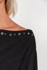 Close-up of a woman wearing a black knit top with silver metal eyelet embellishments along the off-the-shoulder neckline, against a plain light background.