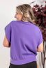 A woman shown from the back wearing a purple short-sleeve knit top, photographed indoors against a light wall with dark red foliage in the background.