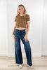 A woman modeling a brown short-sleeve crop top with white polka dots and ruffle trim, paired with dark blue wide-leg jeans and beige boots, standing in a bright room with white walls and light wood floors.