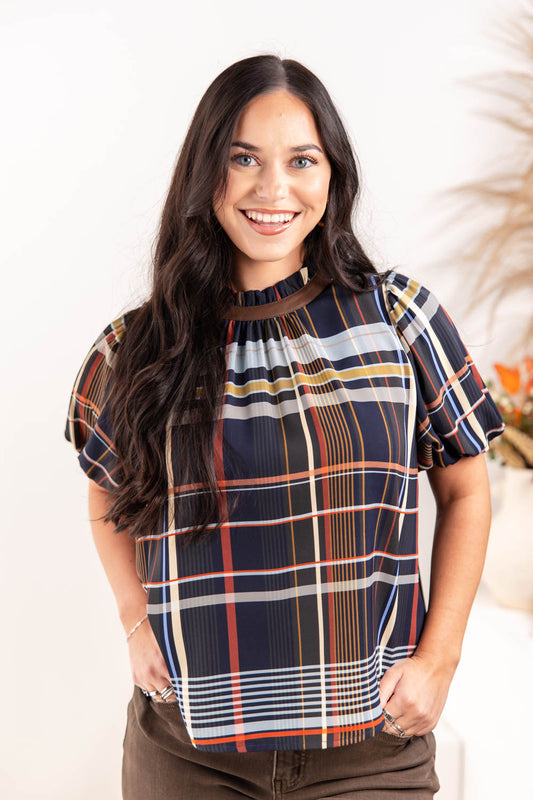 A woman wearing a short-sleeve plaid blouse with navy, orange, white, and yellow stripes, paired with brown pants.