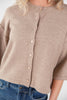 A close-up of a woman wearing a cropped, short-sleeve knit cardigan in a warm taupe-beige color, paired with blue jeans. The cardigan features a round neckline and a front button closure with matching beige buttons.