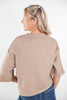 A woman shown from the back wearing a cropped, short-sleeve knit sweater in a light taupe-beige color, paired with blue jeans against a plain white studio background.
