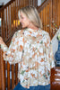 A woman wearing a beige long-sleeve blouse with a white, orange, and green floral pattern, standing in front of a wooden staircase.