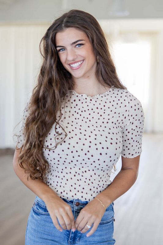 A smiling young woman with long wavy brown hair wearing a cream short-sleeve top with dark brown polka dots and blue jeans, standing in a bright indoor room.