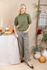 A woman models an olive green short-sleeve mock neck top paired with black and tan gingham wide-leg pants, standing in a styled autumn-themed room with pumpkins, a table, and a ladder.