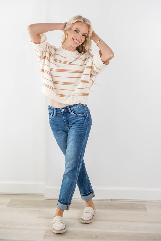 A woman modeling a cream knit sweater with thin tan stripes, paired with blue jeans and cream loafers, standing against a white wall and light wood floor.