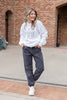 A woman stands outdoors in front of a brick building, wearing a white crewneck sweatshirt with three black embroidered graphic, dark gray joggers, and white sneakers.
