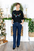 A woman stands wearing a black crewneck sweater with colorful Christmas lights across the chest and dark blue pinstriped wide-leg jeans.