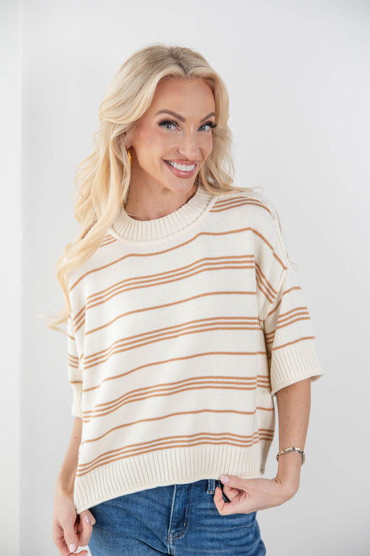 A smiling blonde woman wearing a cream knit sweater with thin horizontal tan stripes and blue jeans, standing against a plain light gray background.