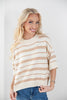 A smiling blonde woman wearing a cream knit sweater with thin horizontal tan stripes and blue jeans, standing against a plain light gray background.