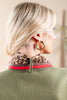 A close-up back and side view of a woman with blonde hair wearing a green knit sweater, a leopard-print neck scarf with a red stripe, and a large gold hoop earring.