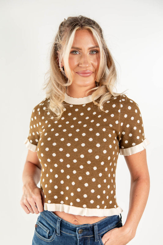 A woman modeling a brown short-sleeve knit top with white polka dots and cream ruffle trim at the collar, sleeve cuffs, and hem, paired with blue jeans against a plain white studio background.