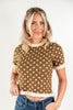 A woman modeling a brown short-sleeve knit top with white polka dots and cream ruffle trim at the collar, sleeve cuffs, and hem, paired with blue jeans against a plain white studio background.