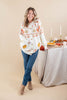 A woman stands in a studio wearing a cream knit cardigan with orange and tan autumn-themed patterns, paired with blue jeans and tan ankle boots. She is posed next to a table decorated with candles, a pie, and fall foliage against a solid beige background.