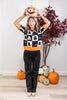 A woman wearing a black and white Halloween sweater with ghost patterns and orange trim, holding a carved pumpkin above her head. She is standing in a decorated room with pumpkins and a potted plant.