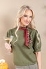 A blonde woman wearing an olive green knit top with a leopard print and red ribbon neck scarf, holding a martini glass against a beige background.