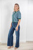 A woman modeling a short-sleeve green and light blue striped knit top with a green collar and cuffs, paired with blue wide-leg jeans and tan shoes, standing in a bright room with white walls and light wood flooring.