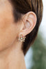 A close-up of a woman's ear wearing a gold snowflake-shaped stud earring with clear rhinestones and small iridescent purple accents.