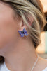 A close-up of a person's ear wearing a bow-shaped earring with purple and white spiderweb patterns and a small black spider in the center, paired with a silver chain necklace.