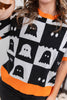A close-up of a person wearing a short-sleeve knit sweater with a black and white ghost checkerboard pattern and bright orange ribbed collar, cuffs, and hem.