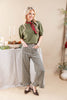 A woman models an olive green short-sleeve top with a red and black patterned neck scarf, paired with green and black houndstooth wide-leg pants with ruffled hems, standing in a styled indoor setting.