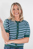 A smiling blonde woman wearing a green and light blue striped short-sleeve knit cardigan with brown buttons, paired with blue jeans, against a plain white studio background.