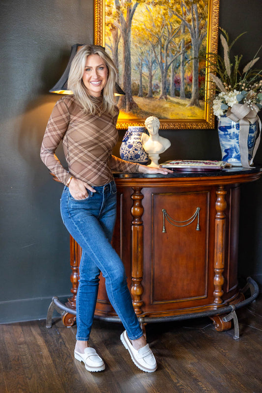 A woman posing in a brown plaid long-sleeve shirt and blue jeans, leaning against a dark wood console table in a decorated room with a painting and a lamp.