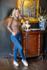 A woman posing in a brown plaid long-sleeve shirt and blue jeans, leaning against a dark wood console table in a decorated room with a painting and a lamp.