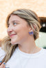 A close-up lifestyle shot of a woman with blonde-highlighted hair wearing a purple bow-shaped stud earring and a delicate silver necklace, looking to the side with a smile.