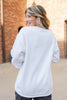 A woman with blonde-highlighted hair is shown from behind wearing a white long-sleeve crewneck sweatshirt in an outdoor setting with a brick building in the background.
