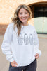 A smiling young woman wearing a white crewneck sweatshirt with a graphic of three simple black ghost outlines, standing outdoors in front of a brick building.