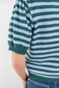 Close-up of a woman wearing a green and white striped knit top with short puff sleeves and a scalloped hem, paired with blue jeans against a plain light background.