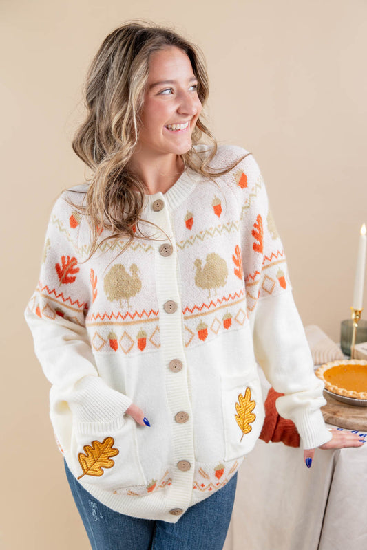 A woman wearing a cream-colored button-up cardigan with autumn-themed knit patterns including turkeys, acorns, and leaves, paired with blue jeans in a warm indoor setting.