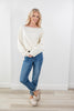 A woman modeling a cream off-the-shoulder knit sweater with silver stud embellishments along the neckline, paired with blue jeans and cream slip-on shoes, standing in a bright room with white walls and light wood floors.
