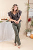 A woman modeling a dark gray patterned ruffled blouse and olive green pants in a warm, neutral-toned indoor setting with a table, candles, and decorative plants.